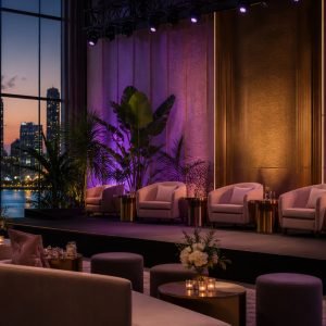 Stage with four beige armchairs and small gold tables, tropical plants, and purple lighting against a city skyline at dusk through large windows.