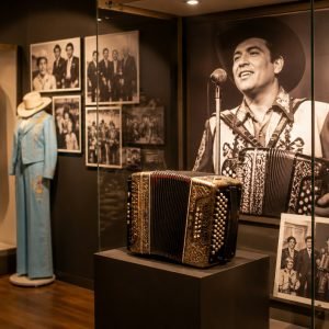 Man in a cowboy hat studies a museum display featuring a vintage accordion, embroidered western outfits, boots, and black-and-white photographs.