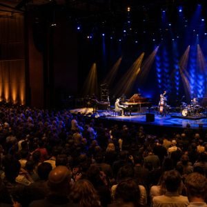 Large audience watching a live band on a dimly lit stage with blue and yellow spotlights, in a packed concert hall.