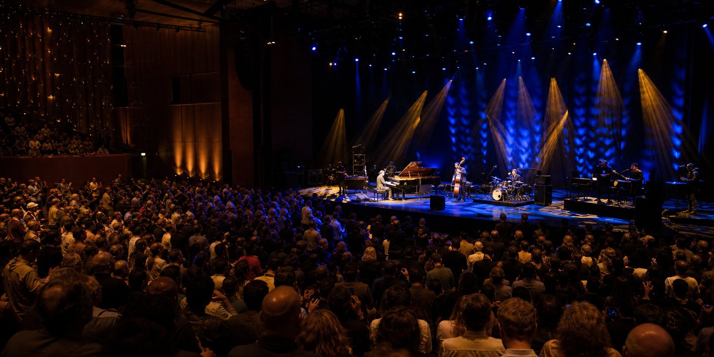 Large audience watching a live band on a dimly lit stage with blue and yellow spotlights, in a packed concert hall.