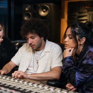Three young adults sit around a large audio mixing console in a recording studio, focused on the controls and discussion of sound.