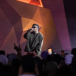 Male singer wearing a dark jacket performs into a microphone on a stage with bold geometric, colorful backdrops; crowd visible in the foreground with phones raised.