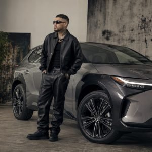 Man in a black leather jacket and sunglasses stands beside a gray modern car in a studio with art and plants in the background, hands in pockets.