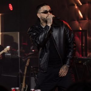 Male singer in a black leather jacket performs on stage with a band, holding a microphone and wearing sunglasses amid red stage lighting.