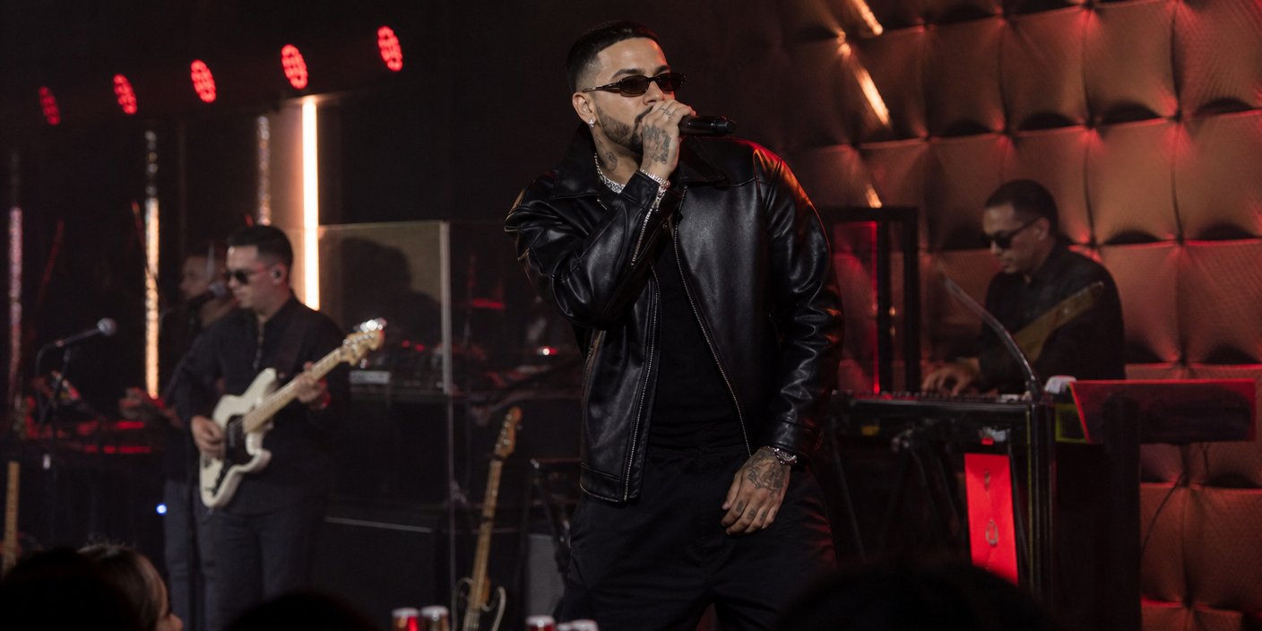 Male singer in a black leather jacket performs on stage with a band, holding a microphone and wearing sunglasses amid red stage lighting.